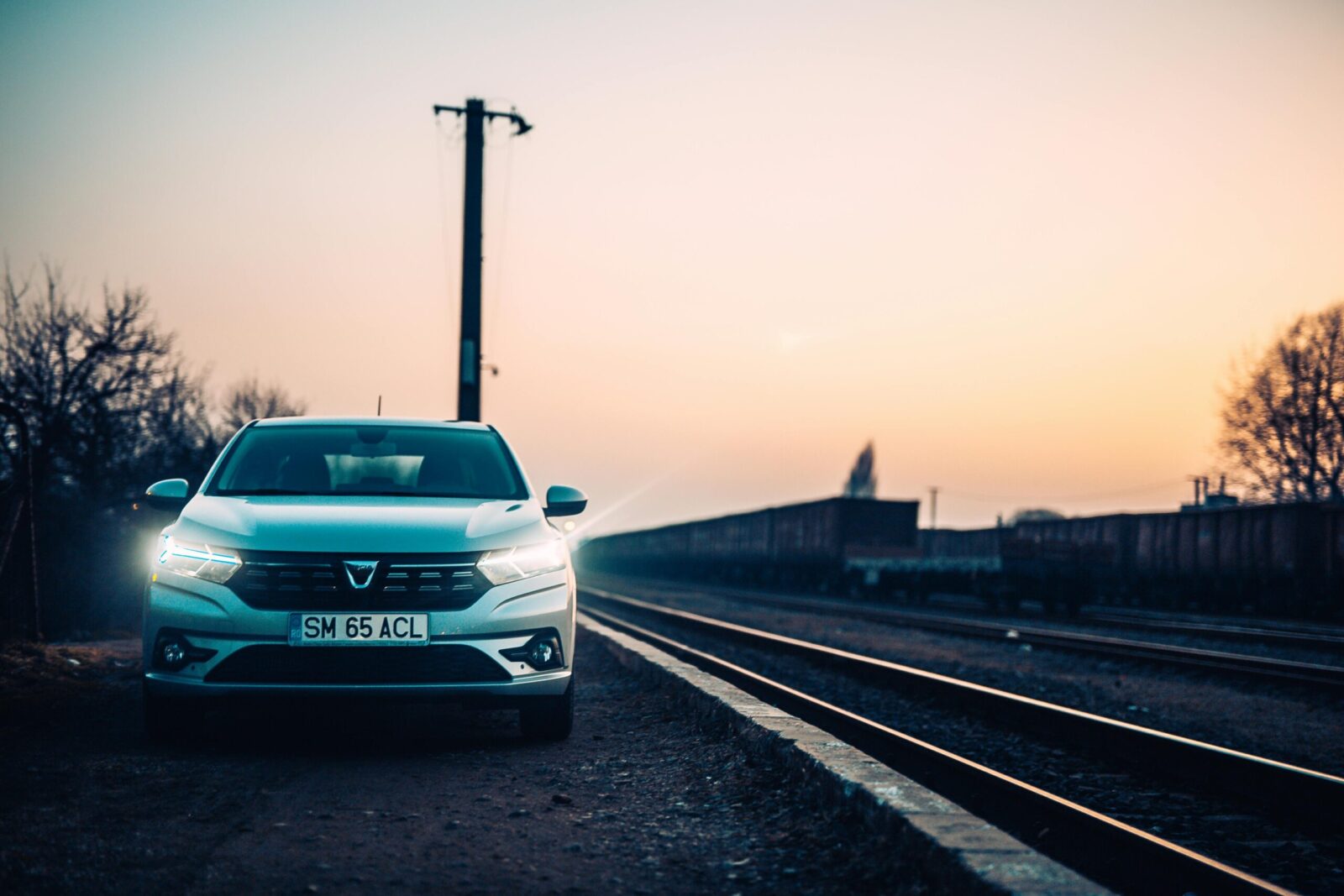 A white car is parked alongside railway tracks during sunset, creating a moody urban scene.